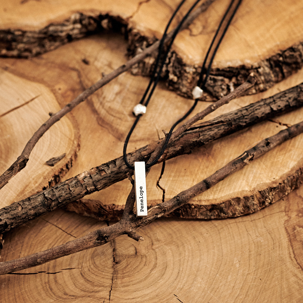Engravable Bar Pendant with a black cord Necklace in Sterling Silver laid down on a log
